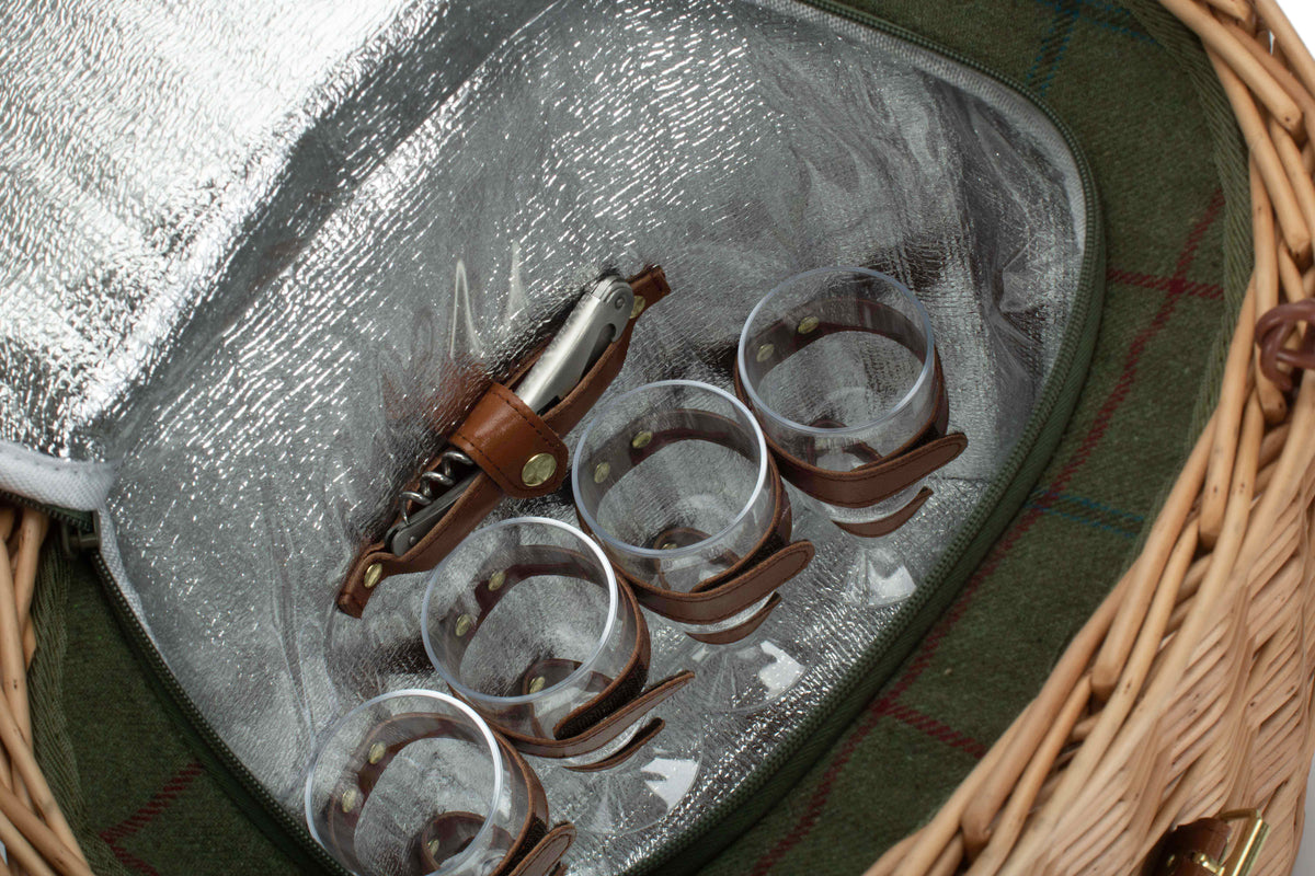 A close-up of a picnic-style wine and glass set inside a wicker basket. The set includes a brown leather-wrapped corkscrew with a metal wine bottle, four clear drinking glasses with brown leather loops, and a silver foil-lined lid.