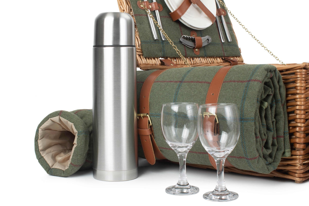 A picnic set including a wicker basket with green plaid fabric lining, a silver thermos bottle, two clear wine glasses, and a rolled green picnic blanket on a white background.