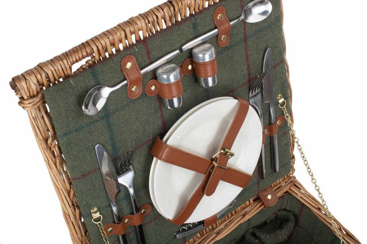 An open wicker picnic basket with a green plaid lining, shown with a white plate, silver cutlery, and brown leather straps on a white background.