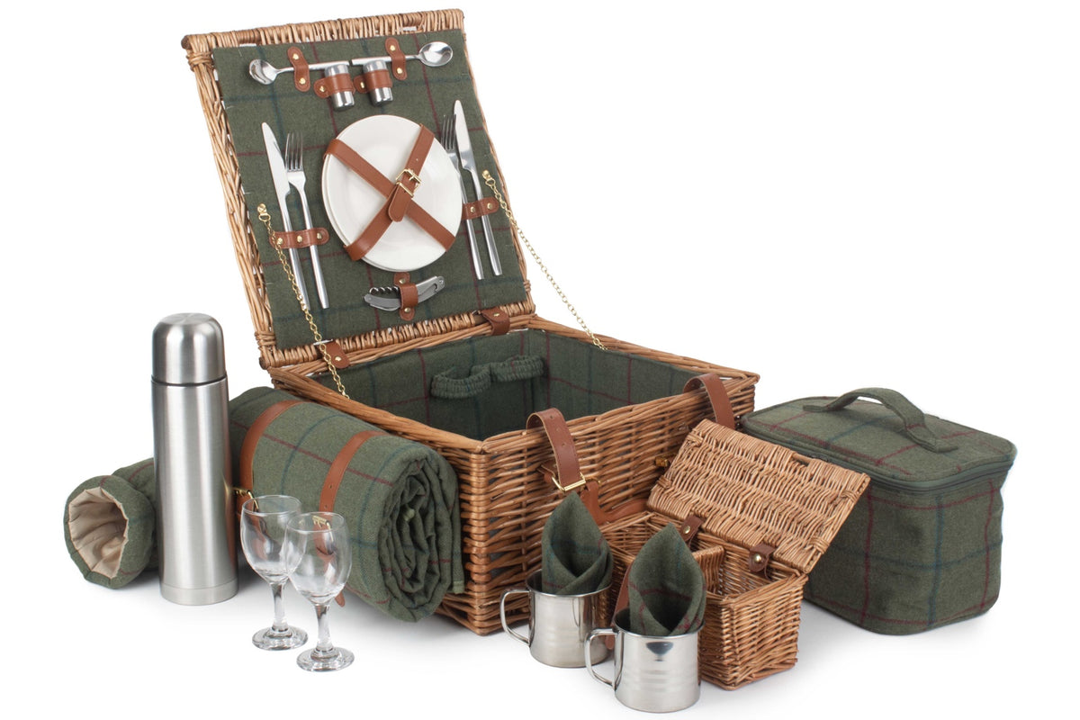A complete picnic basket set displayed on a white background, including a large wicker picnic basket with its lid open, a rolled blanket, a silver thermos, two wine glasses, two metal mugs, and a green fabric bag.